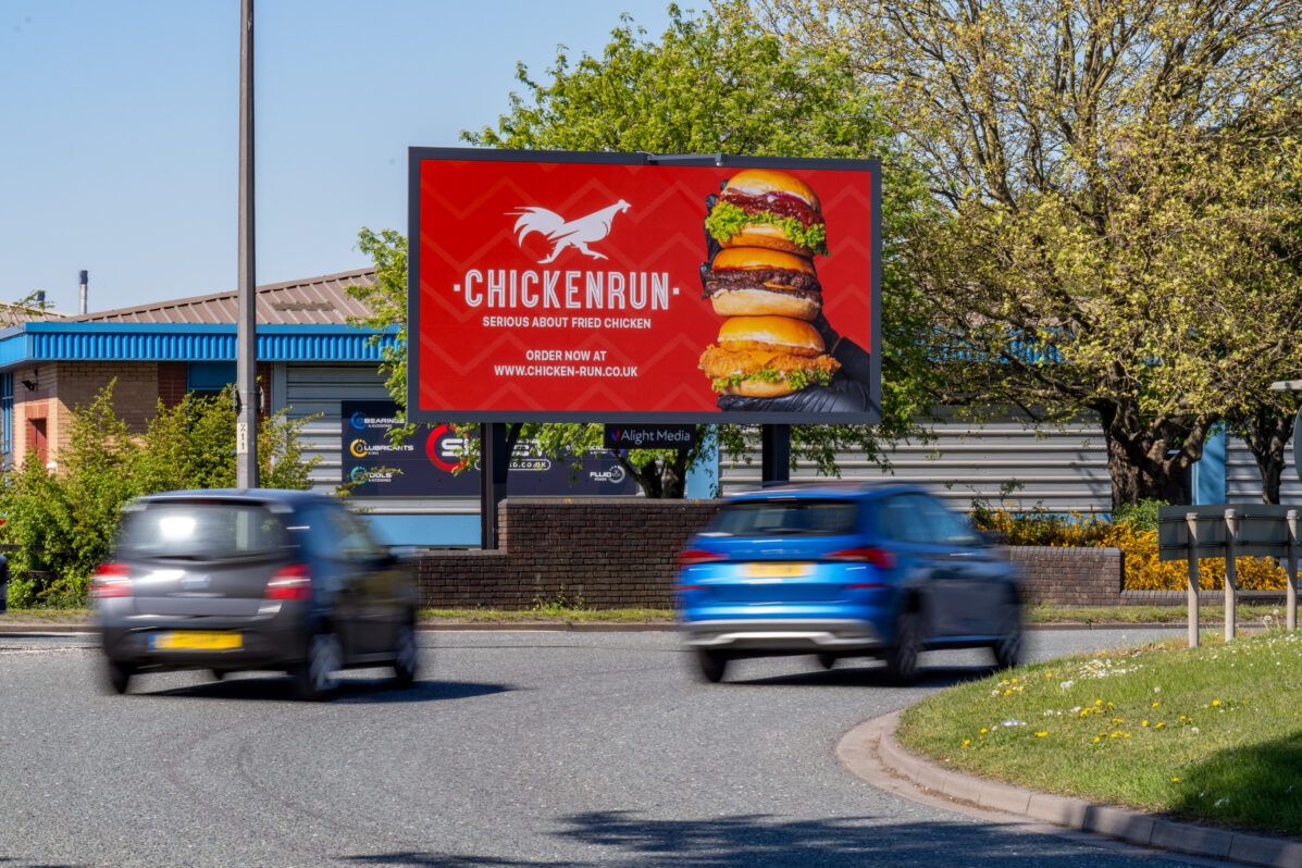 A 48-sheet digital billboard showing a burger ad beside a busy road